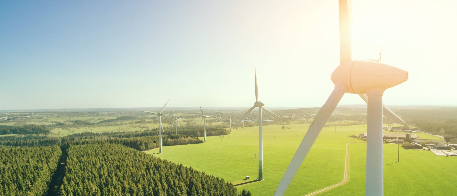 The image depicts a landscape featuring several large wind turbines situated in an open green field. The background reveals a vast expanse of forest and fields under a clear blue sky, suggesting a rural or semi-rural environment. Sunlight is illuminating the scene, creating a sense of bright and pleasant weather conditions, ideal for renewable energy generation from wind power sources. The perspective of the image showcases the height and scale of the wind turbines, highlighting their significance in the field of sustainable energy.