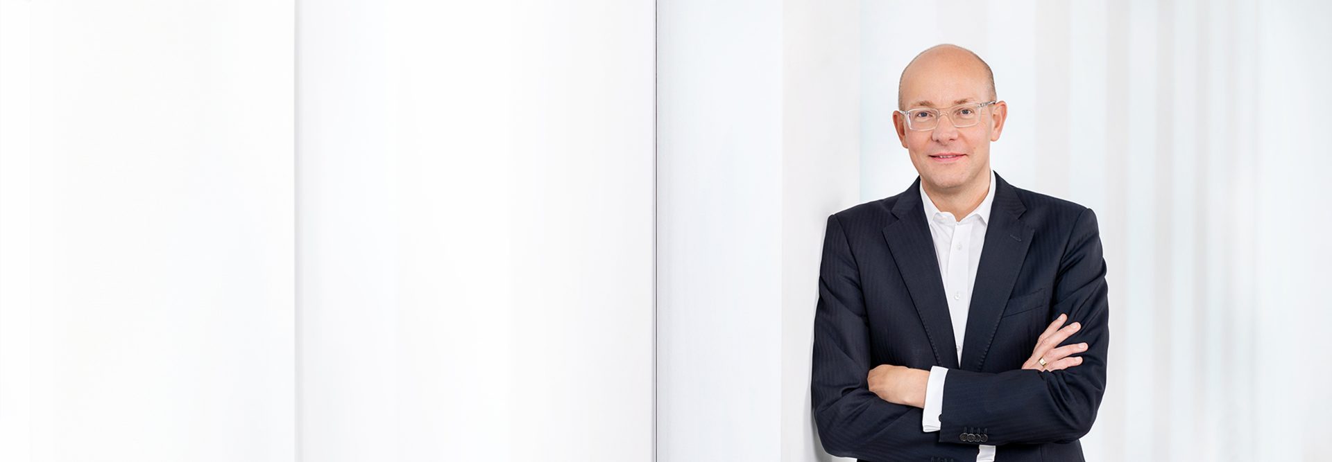 The image shows a person standing indoors with crossed arms, dressed in a formal suit, against a background featuring white columns or vertical panels.